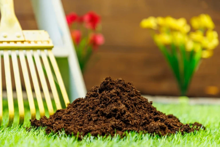 Pile of rich potting soil on artificial grass with a yellow garden rake and blurred spring flowers in background